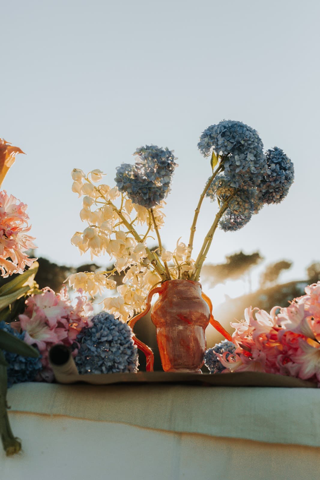 Outdoor tablescape at golden hour with a handmade ceramic vase, blue hydrangea and pale florals
