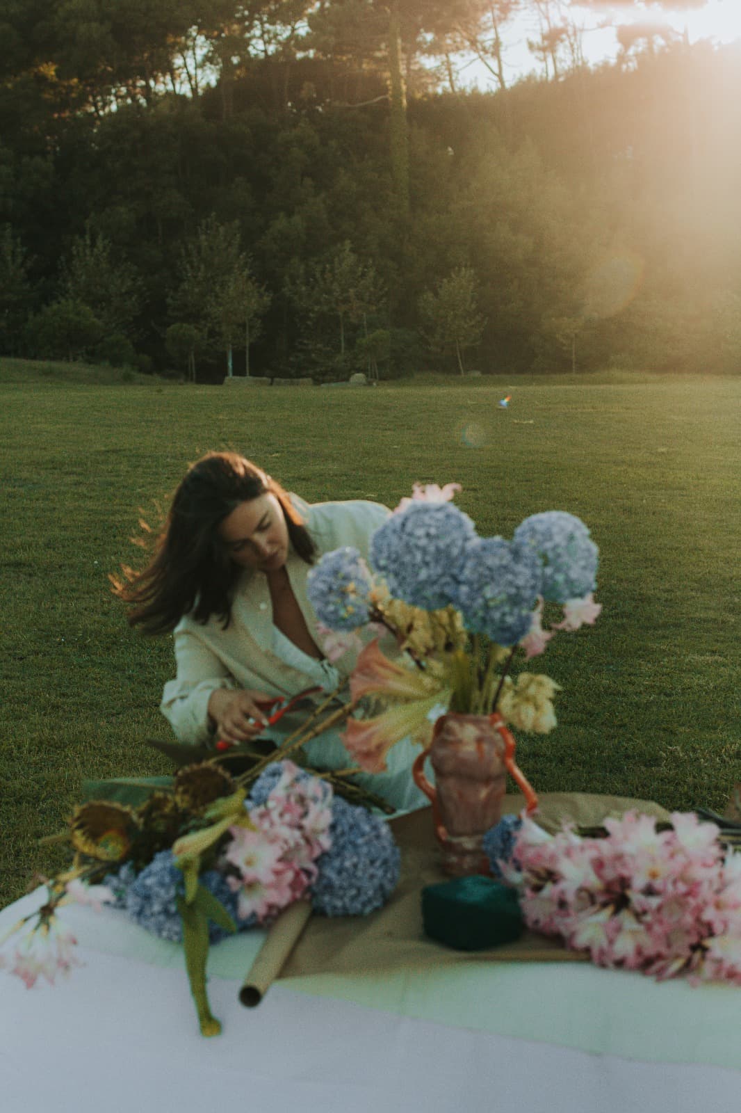 Person kneeling at an outdoor table, arranging flowers with scissors in hand, golden hour