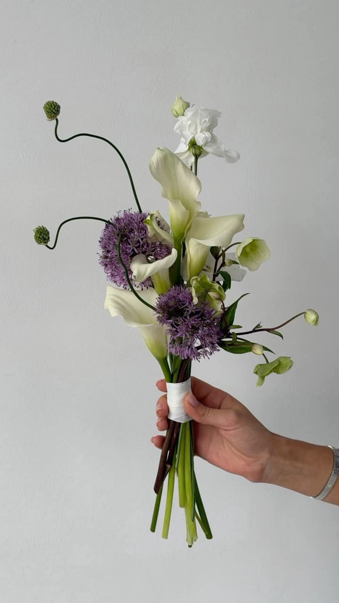 Size S bouquet example: a slender hand-tied bouquet of white calla lilies, purple allium, white stock, and trailing green accents, held in one hand to show its compact scale