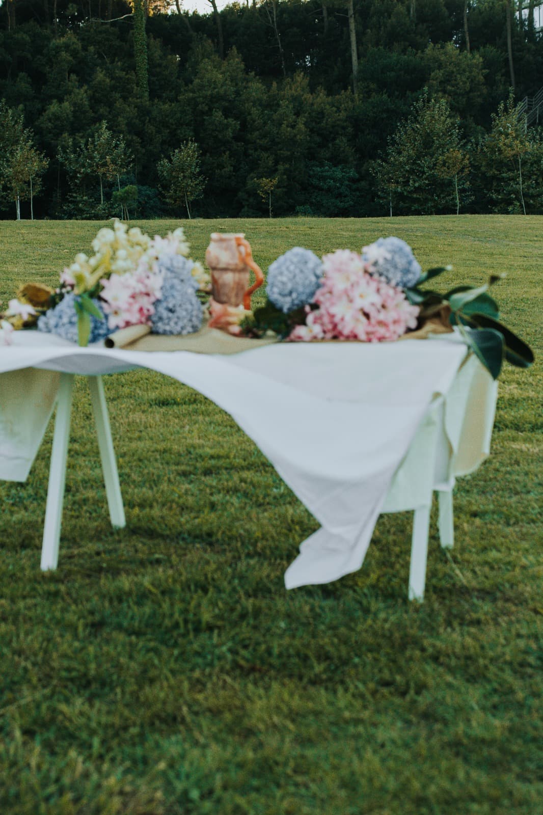 Outdoor tablescape at dusk with a white cloth, blue hydrangea and blush flowers in a terracotta vase